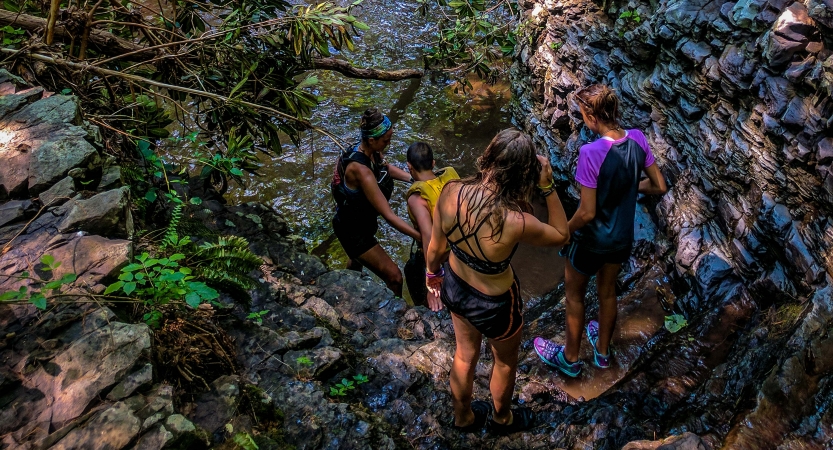 A group of young people wearing swimwear descend a rocky incline toward a body of water. 